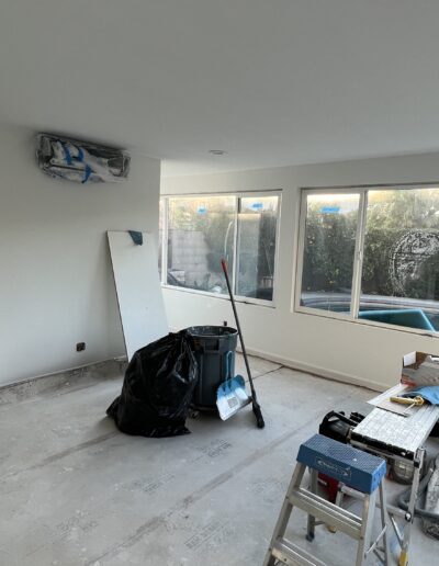 A room under renovation with tools, paint cans, and a ladder. Large windows on two walls provide ample natural light. A doorframe is visible on the left. The floor appears unfinished.