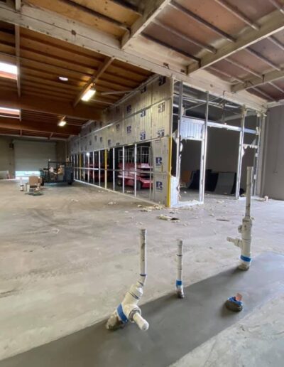 A partially constructed interior with visible plumbing fixtures and an unfinished wall. Construction materials and equipment are scattered across the floor. The roof and ceiling are exposed.