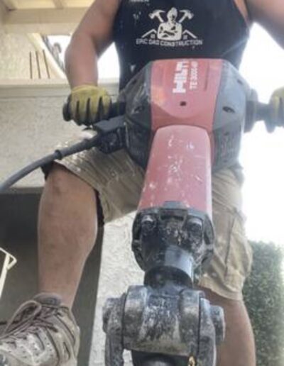 Person wearing a mask, gloves, and a sleeveless shirt operates a concrete breaker, focusing on breaking a concrete surface. House and greenery are visible in the background.