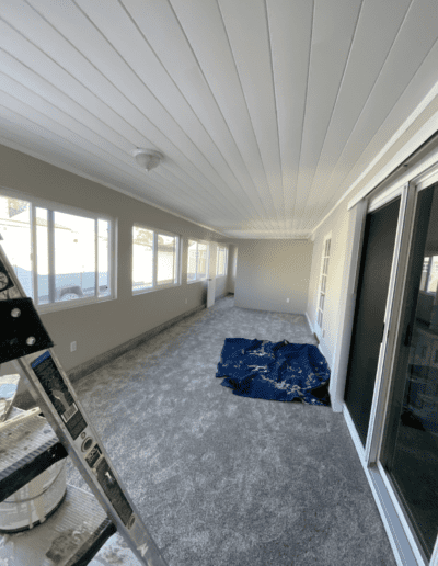 A long room with large windows, gray carpet, and a white ceiling. A ladder with a paint can, a blue tarp on the floor, and sliding glass doors are visible.