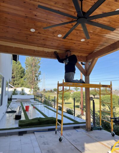 A person stands on scaffolding under a wooden ceiling with a large fan, working with tools. The patio is adjacent to an artificial grass lawn and a low retaining wall. Trees and power lines are visible in the background.