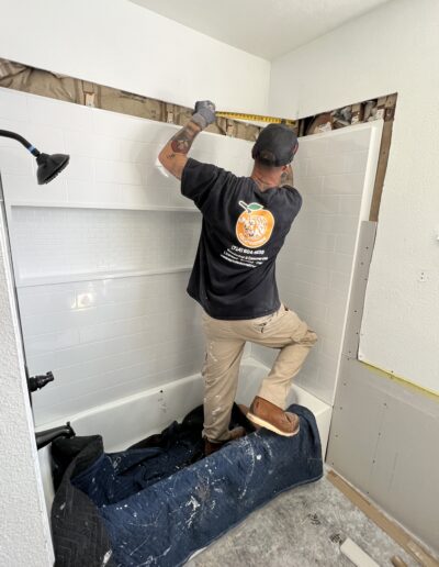 A worker wearing gloves and a cap measures tiles above a bathtub in a bathroom under renovation, with a blue tarp covering part of the tub.