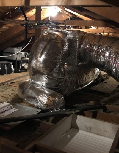 Attic space with exposed wooden beams, flexible ductwork, and various stored items. The area is illuminated by a single lightbulb.