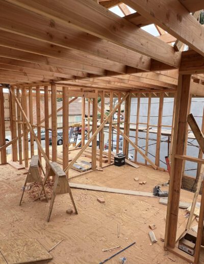 A partially constructed house with wooden framing and beams. Building materials and tools are scattered on the floor. Sunlight filters through various openings in the framework.