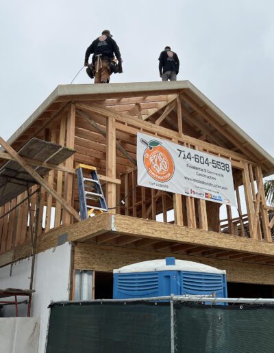 Two workers stand on the roof frame of a partially constructed wooden house. A portable toilet and a banner with a phone number hang in front.