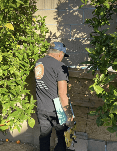 A man in a black cap and t-shirt holds a power tool and a clipboard, working on a wooden barricade in a garden area surrounded by green foliage.