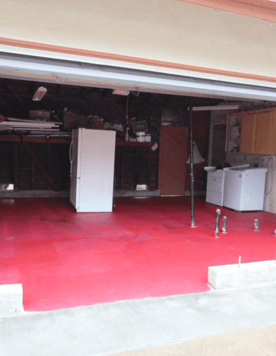 An open garage with a red painted floor, partially finished walls, a white refrigerator, a washing machine, and some pipes visible.
